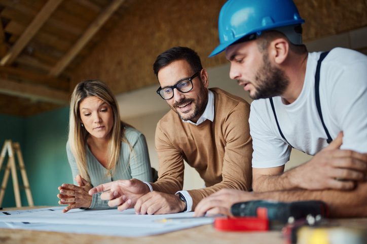 Young couple and their manual worker cooperating while examining housing plan at construction site.